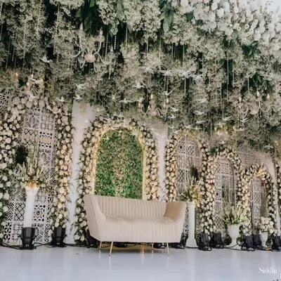 This all-white engagement stage is the definition of timeless elegance. I used a lush green backdrop as a canvas and layered it with thousands of white roses, baby's breath, and hanging floral installations to create a dreamy, ethereal atmosphere. The intricate jaali panels and a mirrored aisle add a touch of modern royalty.