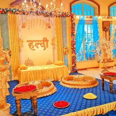A grand Haldi ceremony setup with a royal theme, featuring golden statues, a decorated divan, and traditional brass urlis filled with flower petals.
