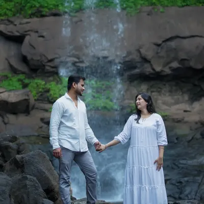 An expecting couple shares a look of love while holding hands, standing on rocks in front of a cascading waterfall. Dressed in coordinated white outfits, this maternity photo has a serene and epic feel, set against a stunning natural landscape.