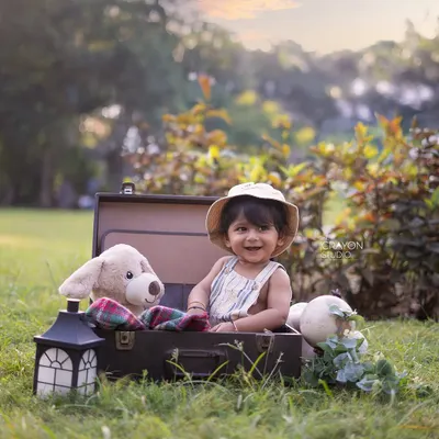 A little explorer is ready for an adventure. This baby boy smiles from inside a vintage suitcase, surrounded by his travel companions in a sunlit park.