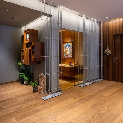 In this apartment foyer, a transparent screen of metal rods separates the entrance from the sacred puja room, creating a visual partition that maintains a sense of openness. The design layers modern materials with traditional art and warm wood flooring.