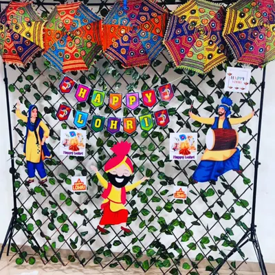 A festive backdrop for a Lohri celebration. The setup includes a "Happy Lohri" banner, traditional Punjabi cutouts, and colorful phulkari-style umbrellas against a green leaf wall.