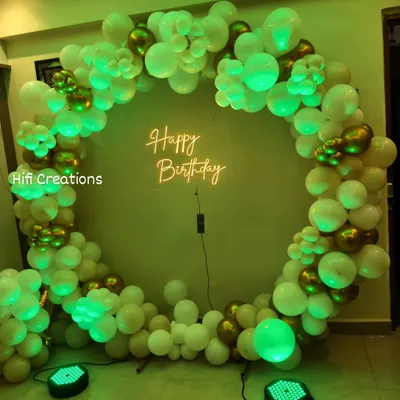A classic and elegant ring backdrop for a birthday celebration. This design uses a full circle of white and chrome gold balloons, lit by a green spotlight, with a 'Happy Birthday' neon sign at the center for a modern and chic look.