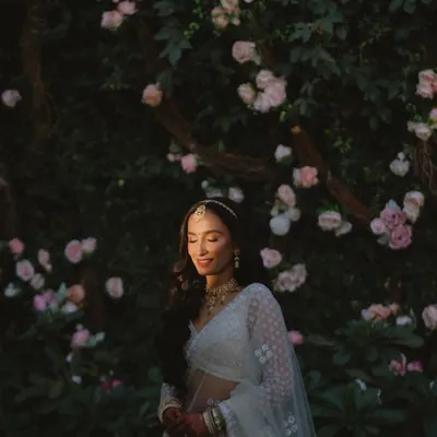 A portrait of a bride bathed in golden light, standing before a wall of blush pink roses. Our floral backdrops are designed to create these soft, romantic, and painterly moments.