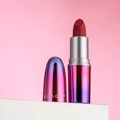 A clean and elegant shot of a Huda Girl lipstick against a soft pink background. The composition is simple, modern, and puts all the focus on the product's unique packaging.
