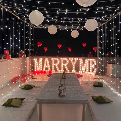 This is another view of a romantic rooftop proposal setup. The 'MARRY ME' marquee letters provide a bold statement, framed by a canopy of fairy lights and paper lanterns for a magical atmosphere.