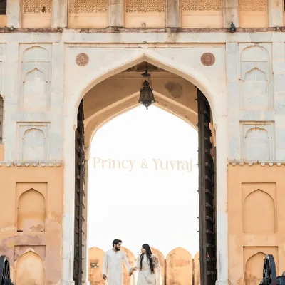 A grand entrance at Amer Fort in Jaipur. The couple, dressed in coordinated white traditional outfits, walks hand-in-hand through the magnificent archway, creating a timeless and regal portrait.