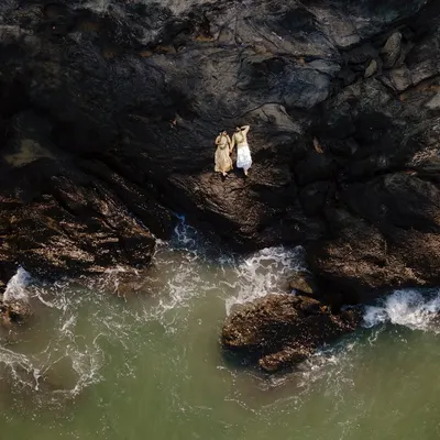 An aerial drone shot captures a couple standing on the edge of dramatic coastal rocks, with green waves washing ashore. This pre-wedding photograph highlights my ability to blend epic landscapes with intimate moments for a truly cinematic feel.