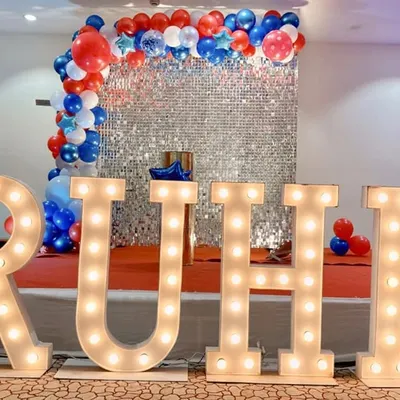 We provide large, illuminated marquee letters to personalize any event. Here, the name 'RUHI' is spelled out in lights, creating a bold and impressive focal point in front of a silver sequin wall and balloon arrangement.