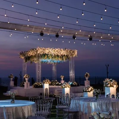 A fairytale sundowner mandap in Goa. This setup features a modern acrylic structure adorned with white florals, set against the purple and orange hues of the sunset, with strings of fairy lights creating a magical ambiance.