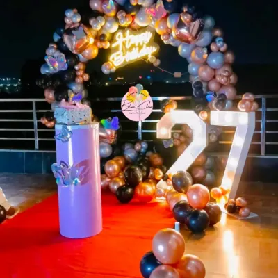 A glamorous 27th birthday party setup on a rooftop, featuring a black, rose gold, and silver balloon arch with a neon sign.