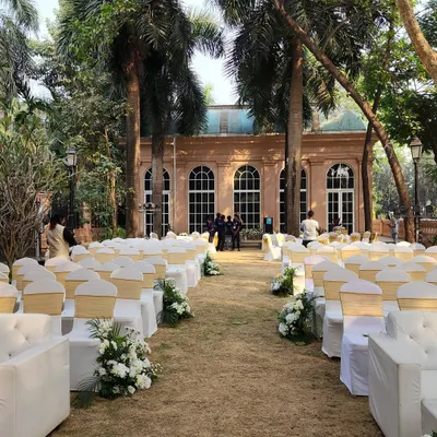 A beautiful outdoor wedding ceremony setup on a lush lawn. The aisle is lined with elegant white guest chairs and floral arrangements, leading towards a classic heritage building that serves as a stunning backdrop.