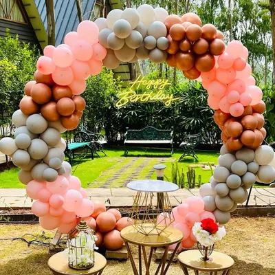 An elegant circular balloon arch in pastel pink, grey, and rose gold, set up outdoors in a garden for a beautiful birthday photo backdrop.