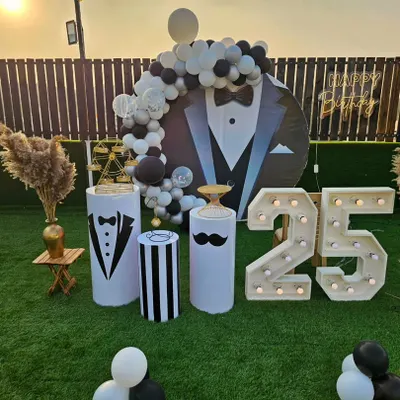 A stylish and fun "tuxedo" theme for a 25th birthday party. The decor includes a custom backdrop, themed pedestals, a balloon arch, and large light-up marquee numbers, all in a classic black and white color scheme.
