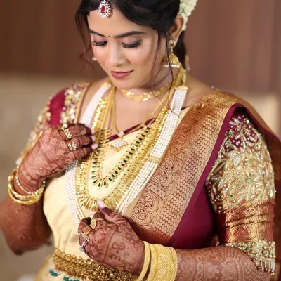 A classic South Indian bridal portrait. The focus is on her intricate gold jewelry and the rich texture of her cream and maroon Kanjeevaram saree, complemented by a soft glam makeup look.