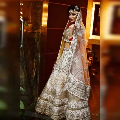 A full-length profile view of the ivory bridal lehenga, showing how the sheer pink dupatta drapes over the shoulder. The maang tikka and bridal jewelry complete the stunning look.