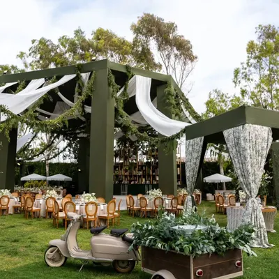 A wide view of a 50th-anniversary garden party I designed. The olive green structure, draped with white fabric and foliage, creates a sophisticated canopy for the celebration, while a vintage scooter adds a touch of whimsical charm.