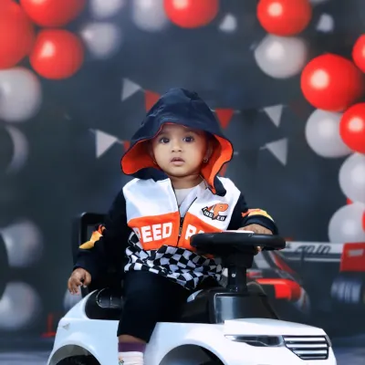 This little racer is ready to hit the track, sitting in a toy car with a cool hooded jacket against a backdrop of red and white balloons.