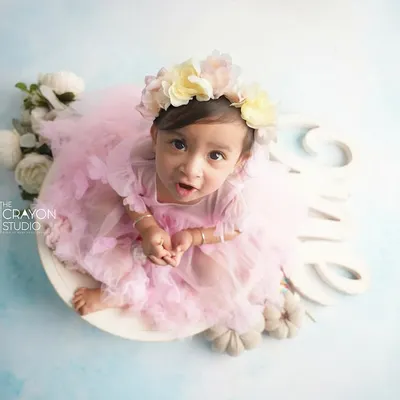 An adorable one-year-old girl in a pink tutu and floral crown, sitting in a white bowl for her first birthday portrait. The "One" sign and soft blue backdrop complete the dreamy look.