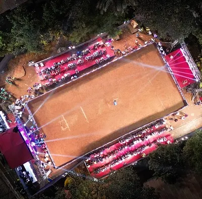 An epic aerial drone shot of the DPL Season 11 underarm cricket tournament at night. This view shows the entire stadium setup, with the brightly lit pitch, spectator stands, and the main stage, all managed by my team.