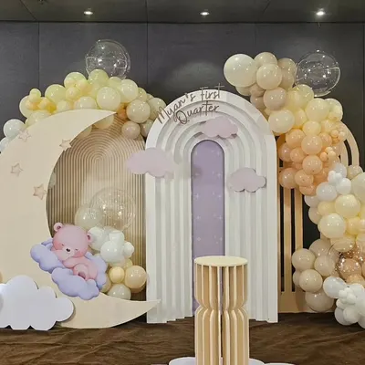 A beautiful celestial-themed setup for "Myan's First Quarter Bash." This decor features a large moon prop with a sleeping teddy bear, a rainbow-arched backdrop, and clouds of soft yellow and white balloons.