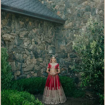 A beautiful portrait of Nita against a rustic stone wall. The contrast between her ornate red lehenga and the natural texture of the wall is striking.