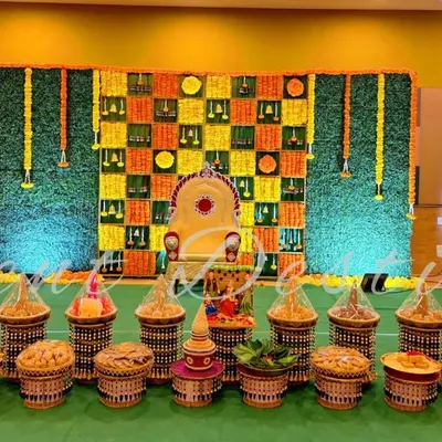 This is a complete setup for a half-saree function, showing both the main stage and the traditional seer plate arrangement. The backdrop features a unique checkerboard pattern of fresh marigolds and green leaves.