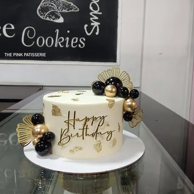 A chic and modern birthday cake with a minimalist aesthetic. The white cake is decorated with black and gold spheres, gold leaf, dried palm fans, and a stylish "Happy Birthday" script topper.
