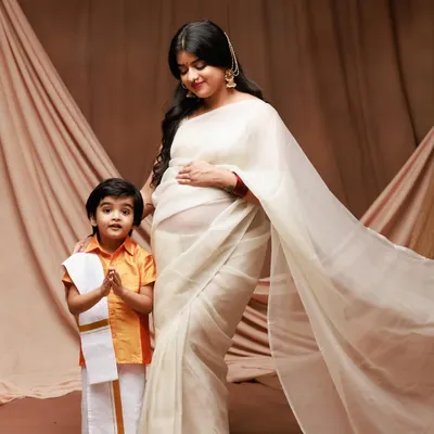 A beautiful maternity portrait celebrating family and tradition. The mother-to-be in a flowing white saree stands with her son, who is dressed in adorable traditional attire.
