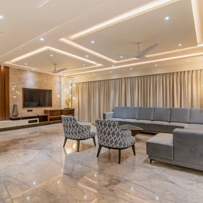 A spacious and bright living room featuring a large sectional sofa, patterned accent chairs, and recessed ceiling lights. The polished marble flooring reflects the light, making the entire area feel open and grand.