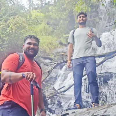 Two trekkers posing in front of a waterfall, fully equipped with backpacks and trekking poles.