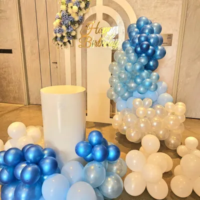 A complete view of the modern decor for a 12th birthday. The setup features a cascading balloon garland in shades of blue and white, a layered arch backdrop, and plinths for the cake and gifts.