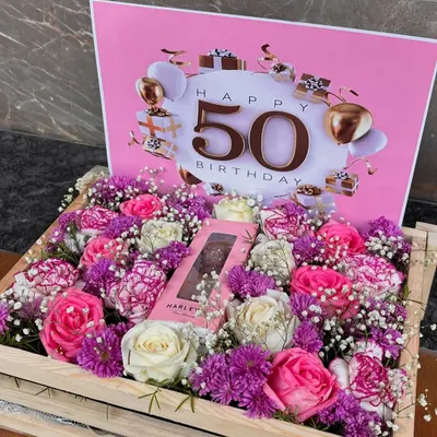 A grand celebration for a milestone. This wooden crate is filled with a vibrant mix of pink and white roses and purple carnations, centered around a chocolate bar and a "Happy 50th Birthday" card.