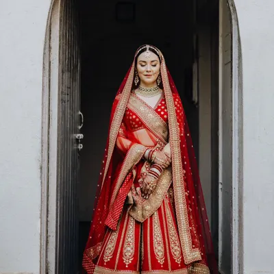 The bride, Rocio, standing in a beautiful archway, looking like a queen in her magnificent red lehenga. A moment of calm elegance before the ceremony.