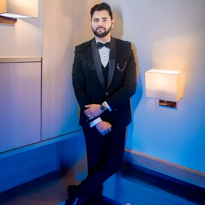 A full-length portrait of the groom, Shubham, looking sharp in his black tuxedo. The cool, blue-toned lighting adds a modern and sophisticated touch.