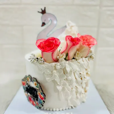 An elegant white cake featuring a graceful swan topper, fresh pink-tipped roses, and delicate lacework-style piping.