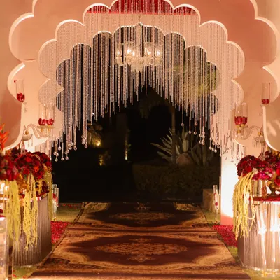 A majestic entrance for a royal-themed wedding, featuring a grand scalloped arch adorned with hanging crystal strings. The path, lined with a traditional patterned carpet and red floral arrangements, exudes opulence.