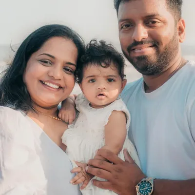 A happy family of three smiling for the camera during their relaxed beach photoshoot. The coordinated white outfits look beautiful against the sea.