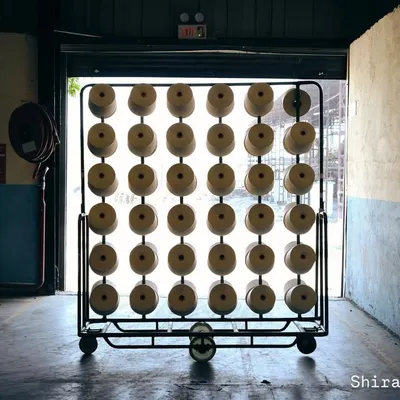 Before the fabric, there is the thread. A rack of yarn cones stands ready in the factory, backlit by the doorway light. This shot captures the quiet, industrial beauty of the manufacturing process.