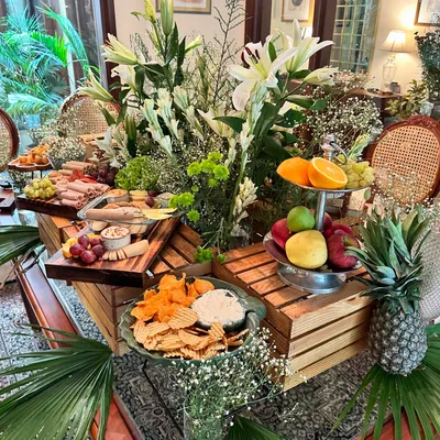 A beautiful grazing table setup for a festive gathering. This display features charcuterie boards, fresh fruit platters, and various dips, all styled with lush palm leaves and white lilies.
