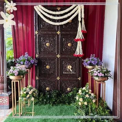 A grand traditional entrance featuring a magnificent carved wooden door, adorned with jasmine garlands. Set against a rich red curtain and surrounded by floral arrangements, this entrance makes a powerful statement.
