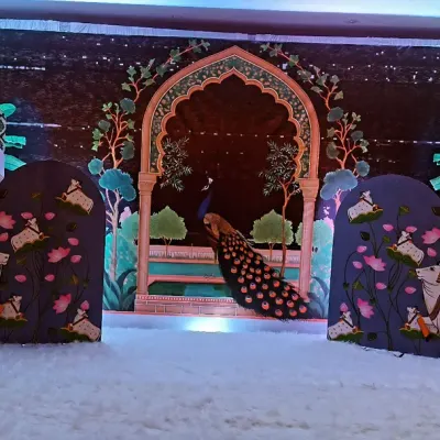 A beautiful stage setup inspired by Pichwai art. This decoration features painted cutouts of cows, lotuses, and a peacock against a night-sky backdrop, creating a scene from Vrindavan.