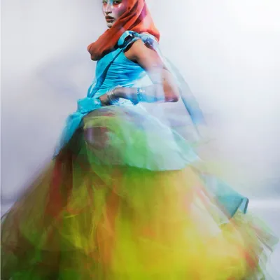 A dynamic shot using long exposure to create a painterly motion blur effect. The model, dressed in a vibrant, multi-colored tulle gown, appears to be in mid-motion, embodying the fluidity and energy of a chemical reaction.