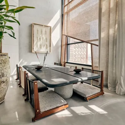 This unique dining setup features low seating and a custom stone-top table, creating a grounded and intimate atmosphere. The design is complemented by natural light filtering through sheer curtains and jali screens.