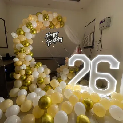 A chic setup for a 26th birthday. This decoration features a half-arch of peach and gold balloons, with large light-up numbers and a "Happy Birthday" neon sign.