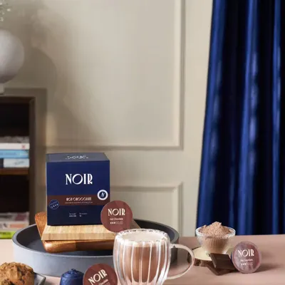 A lifestyle shot for Noir coffee, featuring their hot chocolate pods. The styling with a rich blue curtain and cookies creates a cozy, premium feel for the brand.