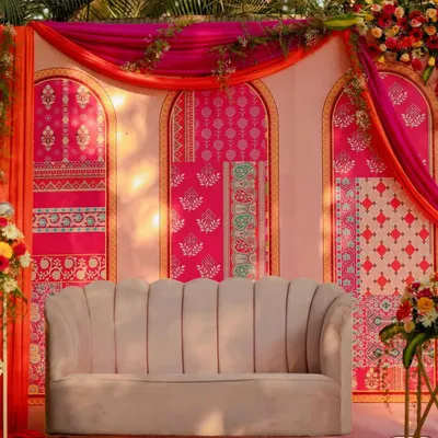 A vibrant Mehendi seating area, featuring a modern scalloped sofa set against a backdrop of colorful block-printed panels and flowing drapes.