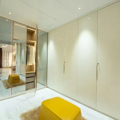 A luxurious walk-in closet concept with high-gloss, floor-to-ceiling wardrobes and a central glass-enclosed island for accessories. A bright yellow ottoman adds a pop of color.