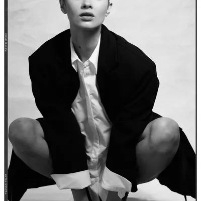 A raw, high-contrast portrait with a minimalist and androgynous feel. The styling consists of an oversized black blazer over a simple white shirt, creating a powerful and confident look with a strong focus on form.