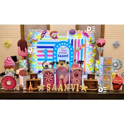 A spectacular Candyland first birthday for Saanvi. This full stage setup is a visual delight with a striped backdrop, giant cupcake and donut props, and a colorful array of balloons.